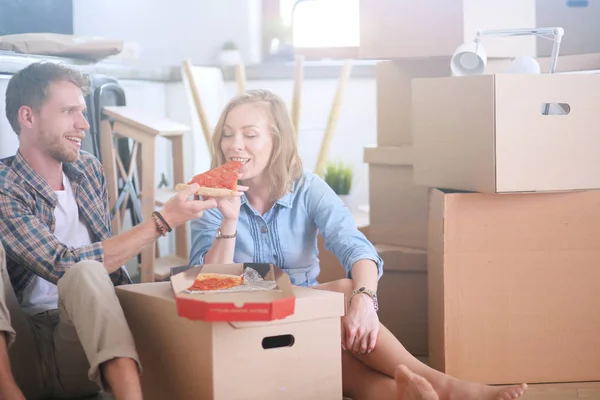 Pareja joven tiene una pausa para comer pizza en el suelo después de mudarse a una nueva casa con cajas a su alrededor. Pareja joven — Foto de Stock