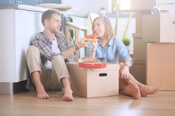 Unga par tar en pizza lunchrast på golvet efter att ha flyttat in i ett nytt hem med lådor runt dem. Unga par — Stockfoto