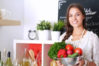Mutfakta elinde sebzelerle gülümseyen genç bir kadın.
