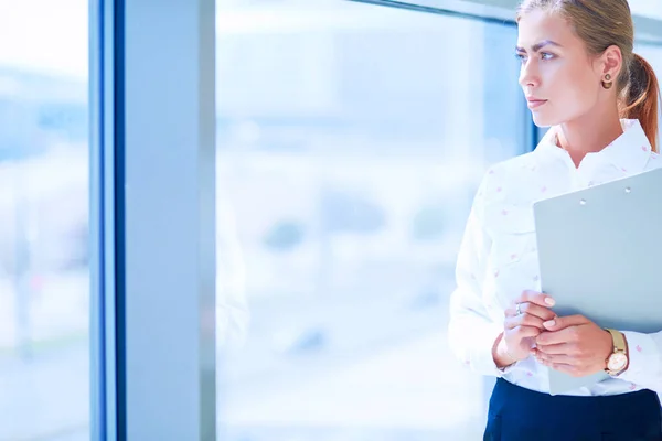 Mujer de negocios de pie contra la ventana de la oficina celebración de ordenador portátil — Foto de Stock