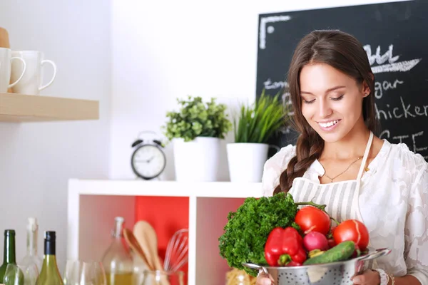 Mutfakta elinde sebzelerle gülümseyen genç bir kadın.
