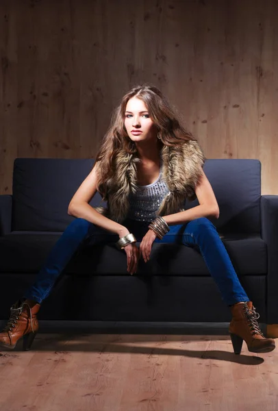 Retrato de una mujer elegante sentada en un sofá negro con pantalones vaqueros azules y chaleco de piel — Foto de Stock