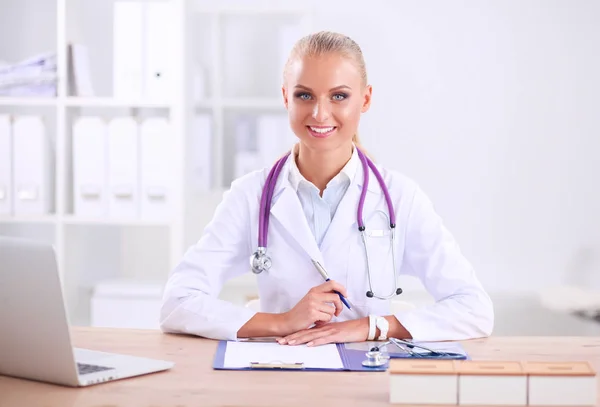 Hermosa joven sonriente doctora sentada en el escritorio y escribiendo — Foto de Stock