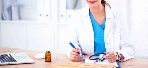 Schöne junge lächelnde Ärztin sitzt am Schreibtisch und schreibt. — Stockfoto