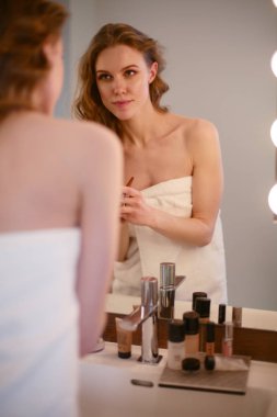 Genç bir kadın aynaya bakıyor ve makyaj yapıyor..