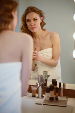 Genç bir kadın aynaya bakıyor ve makyaj yapıyor..
