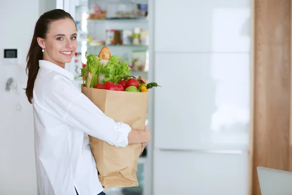 Mujer joven sosteniendo bolsa de la compra de comestibles con verduras.De pie en la cocina. Mujer en la cocina mirando a la cámara — Foto de Stock