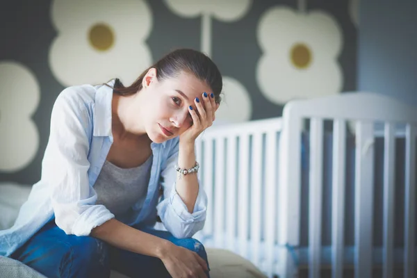 Ung trött kvinna som sitter på sängen nära barnens spjälsäng. Ung mamma. — Stockfoto