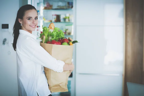 Jeune femme tenant sac d'épicerie avec des légumes. Debout dans la cuisine. Femme dans la cuisine regardant la caméra — Photo