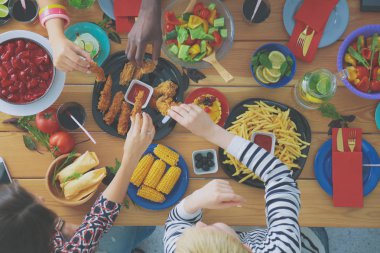 Ahşap masada otururken birlikte yemek yiyen bir grup insanın üst düzey görüntüsü. Masada yemek var. İnsanlar fast food yiyor..