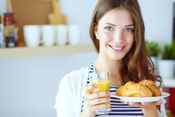 Mujer joven con vaso de jugo y pasteles de pie en la cocina —  Fotos de Stock