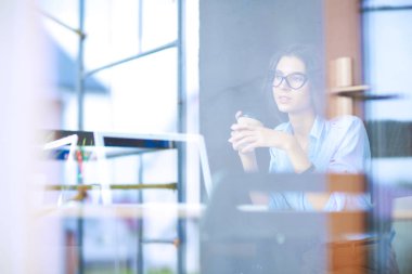 Ofis masasında dizüstü bilgisayarıyla oturan genç bir kadın. Pencereden bakıyor. Genç kadın.