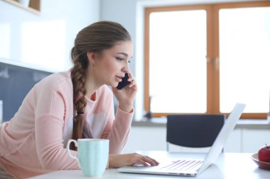 Genç kadın bir dizüstü bilgisayar kullanarak ve cep telefonuyla konuşurken mutfak masasında oturur.