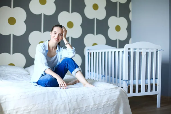 Junge Frau, die auf dem Bett neben dem Kinderbett sitzt. junge Mutter — Stockfoto