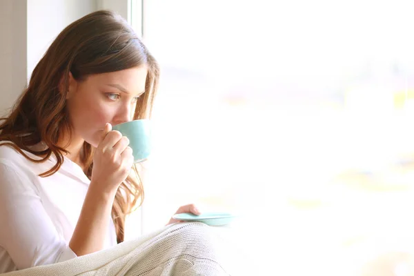 Giovane donna a casa seduta vicino alla finestra rilassarsi nel suo soggiorno lettura libro e bere caffè o tè. Giovane donna a casa — Foto Stock