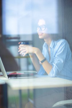 Ofis masasında dizüstü bilgisayarıyla oturan genç bir kadın. Pencereden bakıyor. Genç kadın.