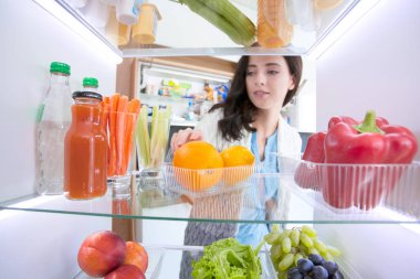 Sağlıklı yiyecek, sebze ve meyvelerle dolu açık buzdolabının yanında duran kadın portresi.