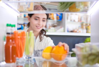 Sağlıklı yiyecek, sebze ve meyvelerle dolu açık buzdolabının yanında duran kadın portresi.