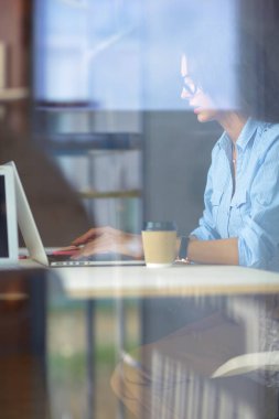 Ofis masasında dizüstü bilgisayarıyla oturan genç bir kadın. Pencereden bakıyor. Genç kadın.