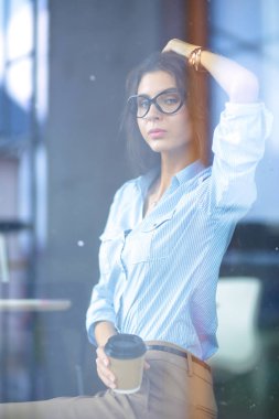 Ofis masasında dizüstü bilgisayarıyla oturan genç bir kadın. Pencereden bakıyor. Genç kadın.