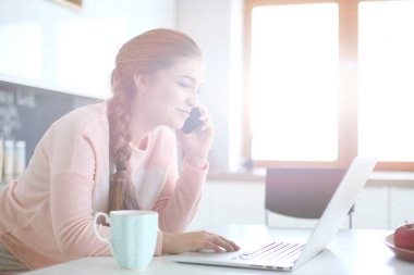 Genç kadın bir dizüstü bilgisayar kullanarak ve cep telefonuyla konuşurken mutfak masasında oturur.