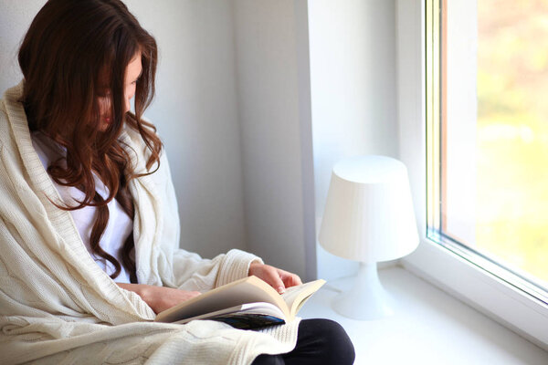Young woman at home sitting near window relaxing in her living room reading book and drinking coffee or tea . Young woman at home