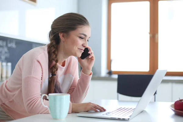 Ung kvinna sitter vid köksbordet med en laptop och talar i en mobiltelefon. — Stockfoto