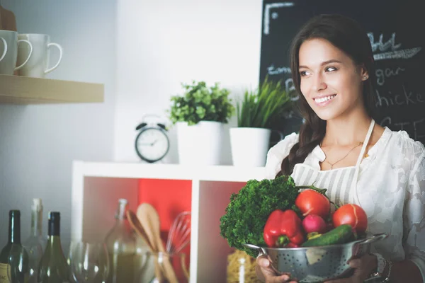 Mujer joven sonriente sosteniendo verduras de pie en la cocina —  Fotos de Stock