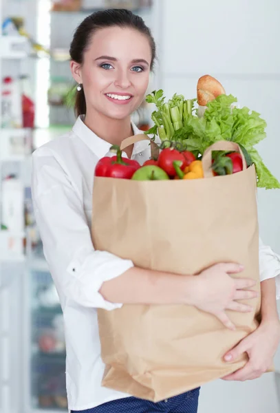 Fiatal nő kezében élelmiszer bevásárló táska zöldségekkel.Álló a konyhában. Egy nő a konyhában a kamerába néz. — Stock Fotó