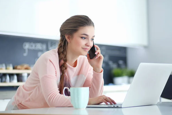 Junge Frau sitzt mit Laptop am Küchentisch und telefoniert. — Stockfoto