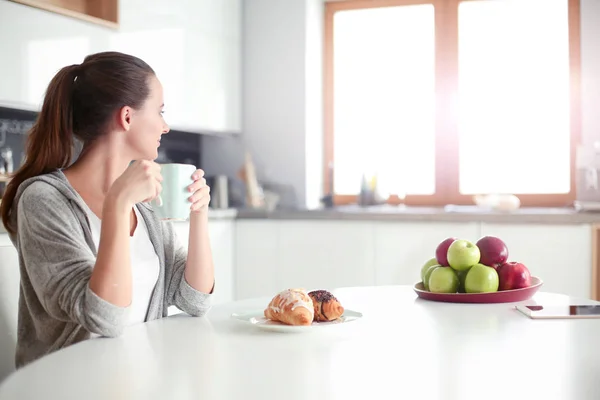 Женщина пьет чай со сладким круассаном за кухонным столом . — стоковое фото