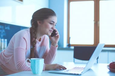 Genç kadın bir dizüstü bilgisayar kullanarak ve cep telefonuyla konuşurken mutfak masasında oturur.
