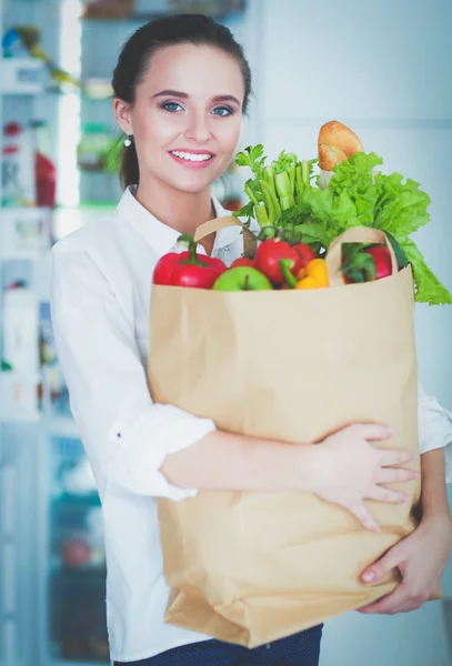 Mujer joven sosteniendo bolsa de la compra de comestibles con verduras.De pie en la cocina. Mujer en la cocina mirando a la cámara — Foto de Stock