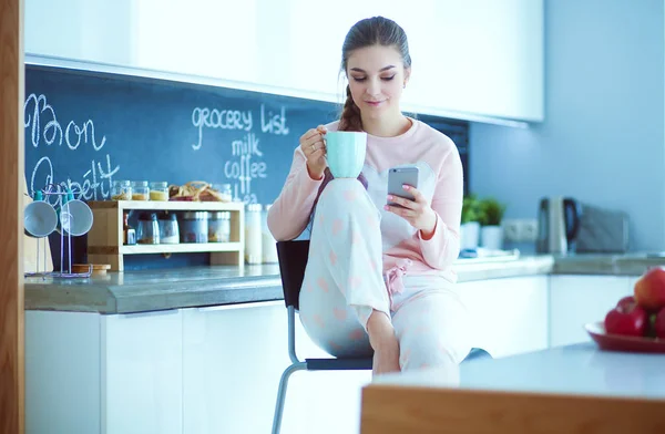 Mujer usando el teléfono móvil sentado en la cocina moderna . — Foto de Stock