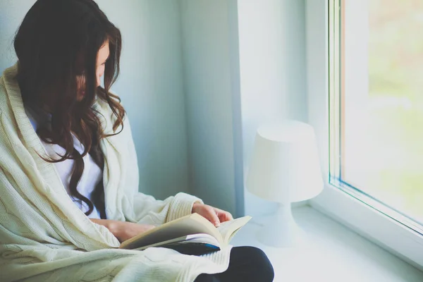 Young woman at home sitting near window relaxing in her living room reading book and drinking coffee or tea . Young woman at home — Stock Photo, Image