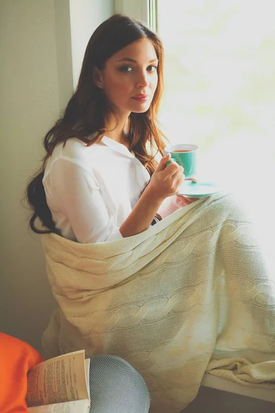 Genç kadın evde oturma odası okuma kitabında rahatlatıcı ve kahve veya çay içme pencere oturuyor. Genç kadın evde