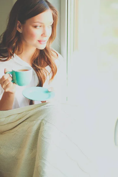 Young woman at home sitting near window relaxing in her living room reading book and drinking coffee or tea . Young woman at home — Stock Photo, Image