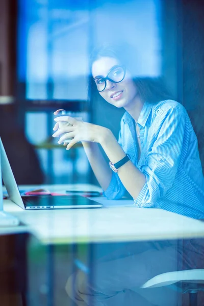 Ofis masasında dizüstü bilgisayarıyla oturan genç bir kadın. Pencereden bakıyor. Genç kadın.