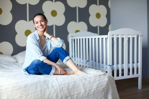 Jonge vrouw zittend op het bed in de buurt van childrens kinderbed. Jonge moeder — Stockfoto