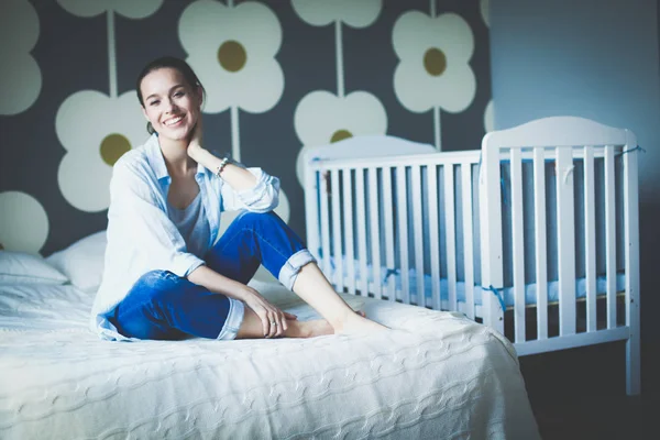 Jonge vrouw zittend op het bed in de buurt van childrens kinderbed. Jonge moeder — Stockfoto