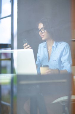 Ofis masasında dizüstü bilgisayarıyla oturan genç bir kadın. Pencereden bakıyor. Genç kadın.