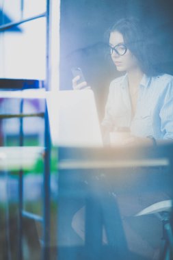 Ofis masasında dizüstü bilgisayarıyla oturan genç bir kadın. Pencereden bakıyor. Genç kadın.