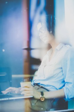 Ofis masasında dizüstü bilgisayarıyla oturan genç bir kadın. Pencereden bakıyor. Genç kadın.