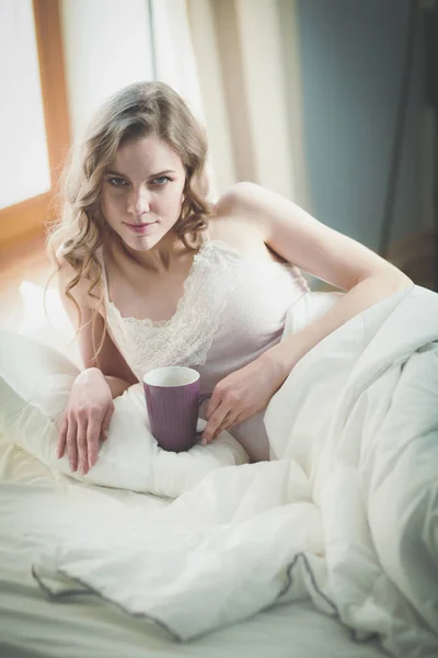 Young woman drinking cup of coffee or tea while lying in bed. — Stock Photo, Image