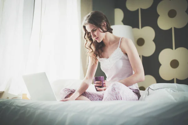 Dizüstü bilgisayar ile yatakta oturan genç güzel kadın. — Stok fotoğraf