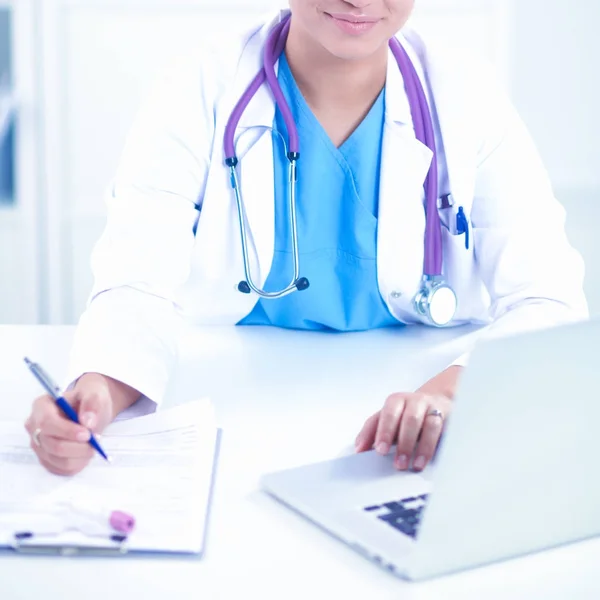 Mooie jonge glimlachende vrouwelijke arts zitten aan het bureau en schrijven. — Stockfoto