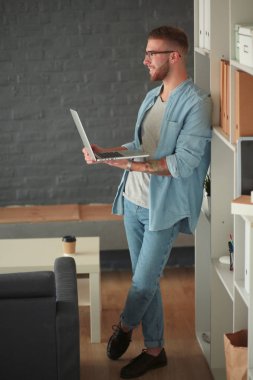 Duvarın yanında dizüstü bilgisayar tutan genç adam..