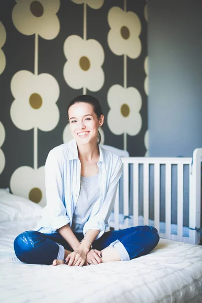 Jonge vrouw zittend op het bed in de buurt van childrens kinderbed. Jonge moeder — Stockfoto