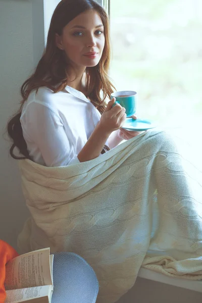 Jovem mulher em casa sentado perto da janela relaxante em sua sala de estar leitura livro e beber café ou chá. Jovem mulher em casa — Fotografia de Stock
