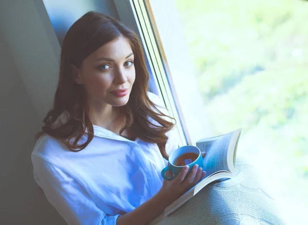 年轻女子在家里坐在靠近窗口轻松的在她的客厅里看书、 喝咖啡或茶。年轻女子在家里 — 图库照片